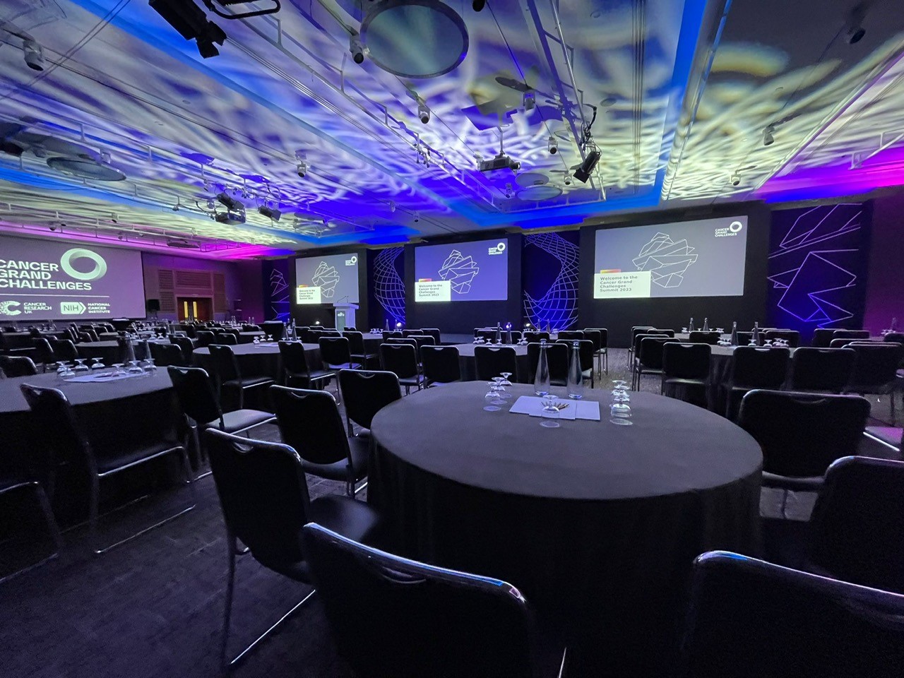 A conference room with a wide stage, screens and ceiling lighting